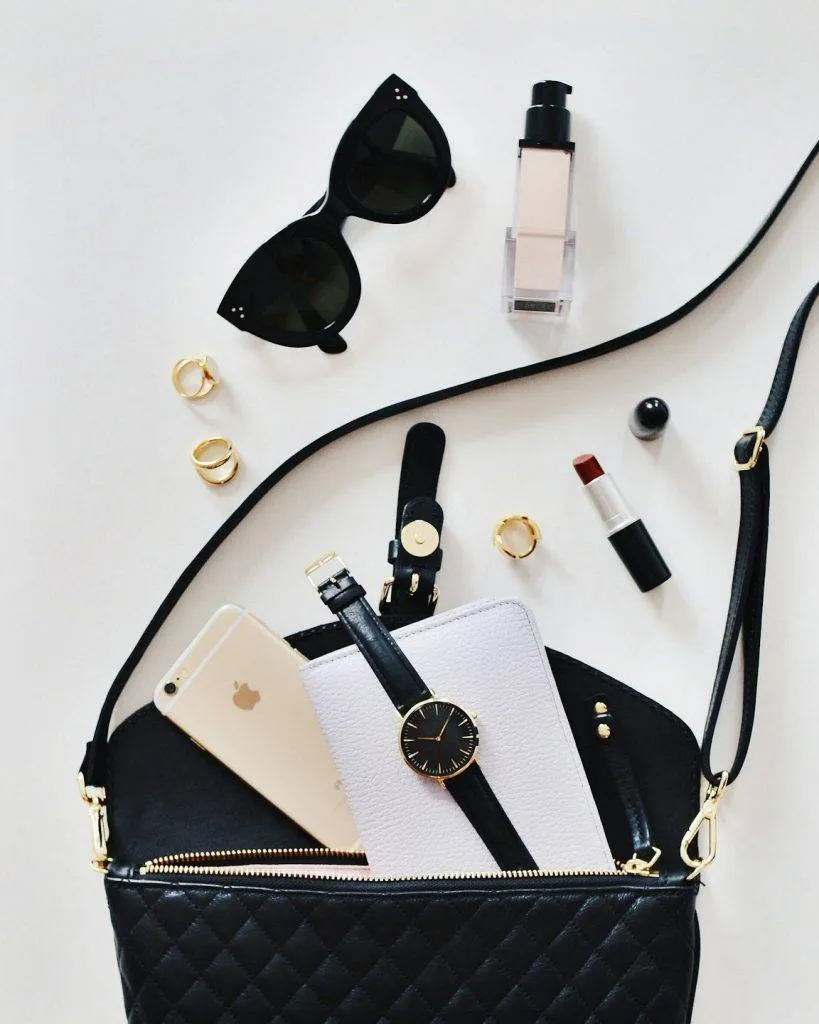 Assorted stylish accessories including a handbag, sunglasses, perfume, watch and smartphone laid out on a white surface